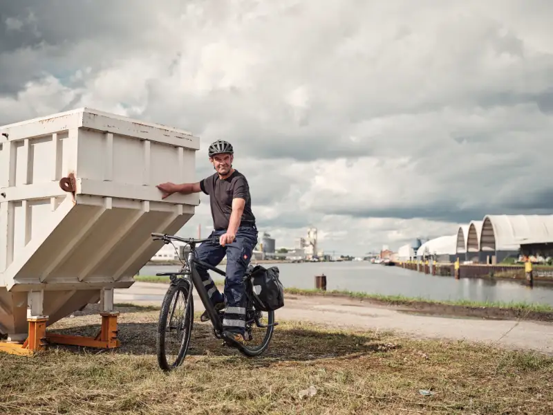 JobRadler in Arbeitshose lehnt an Container, hinter ihm Wasser